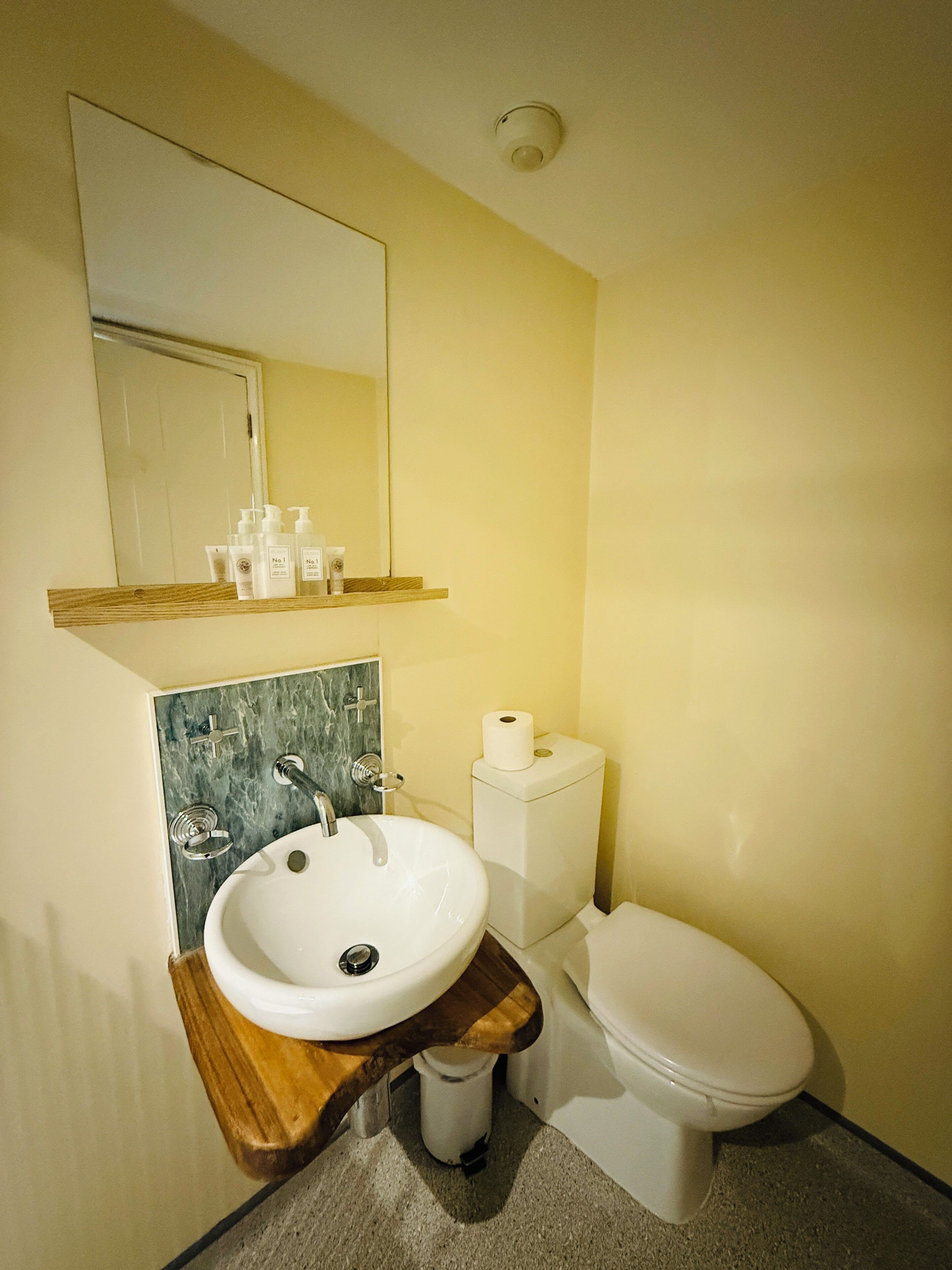 A bathroom with a wooden vanity, a white sink, a mirror, a toilet, and a shelf with toiletries.