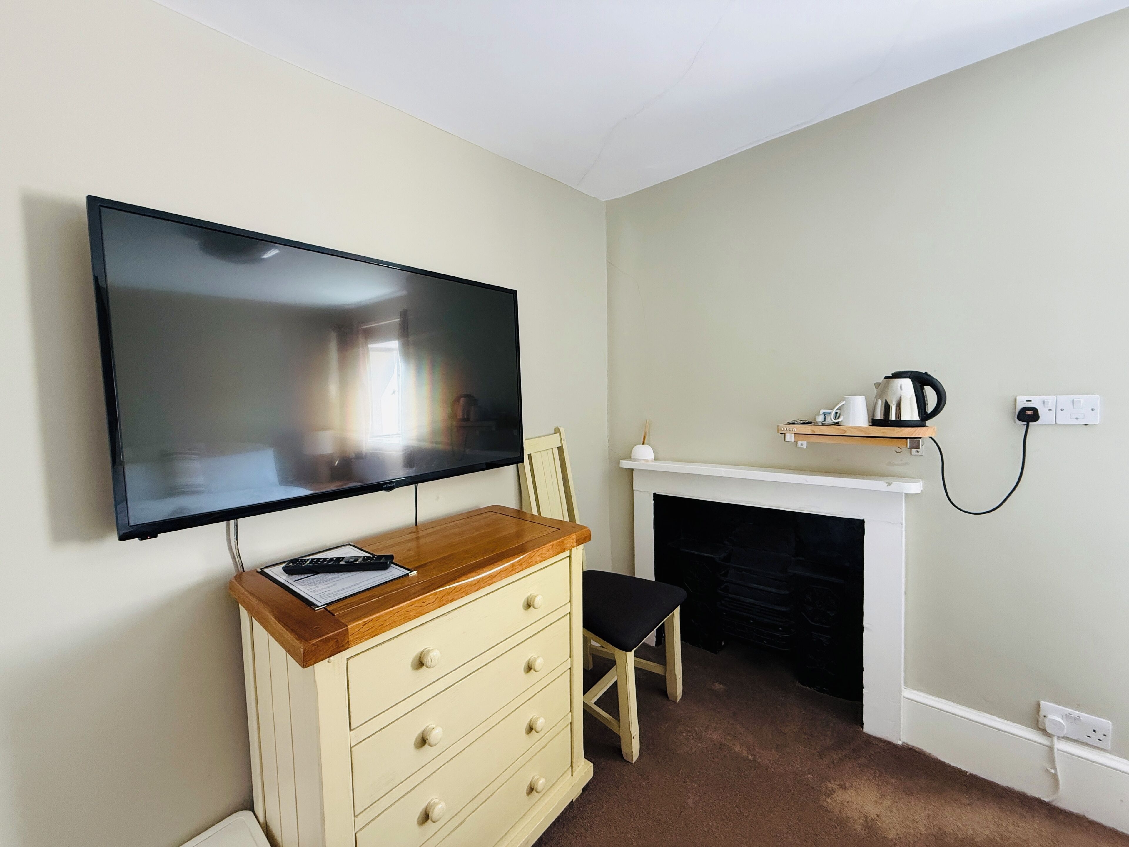 A room with a flat-screen TV mounted on the wall, a wooden dresser with drawers, a black chair, a white fireplace, and a shelf above the fireplace.