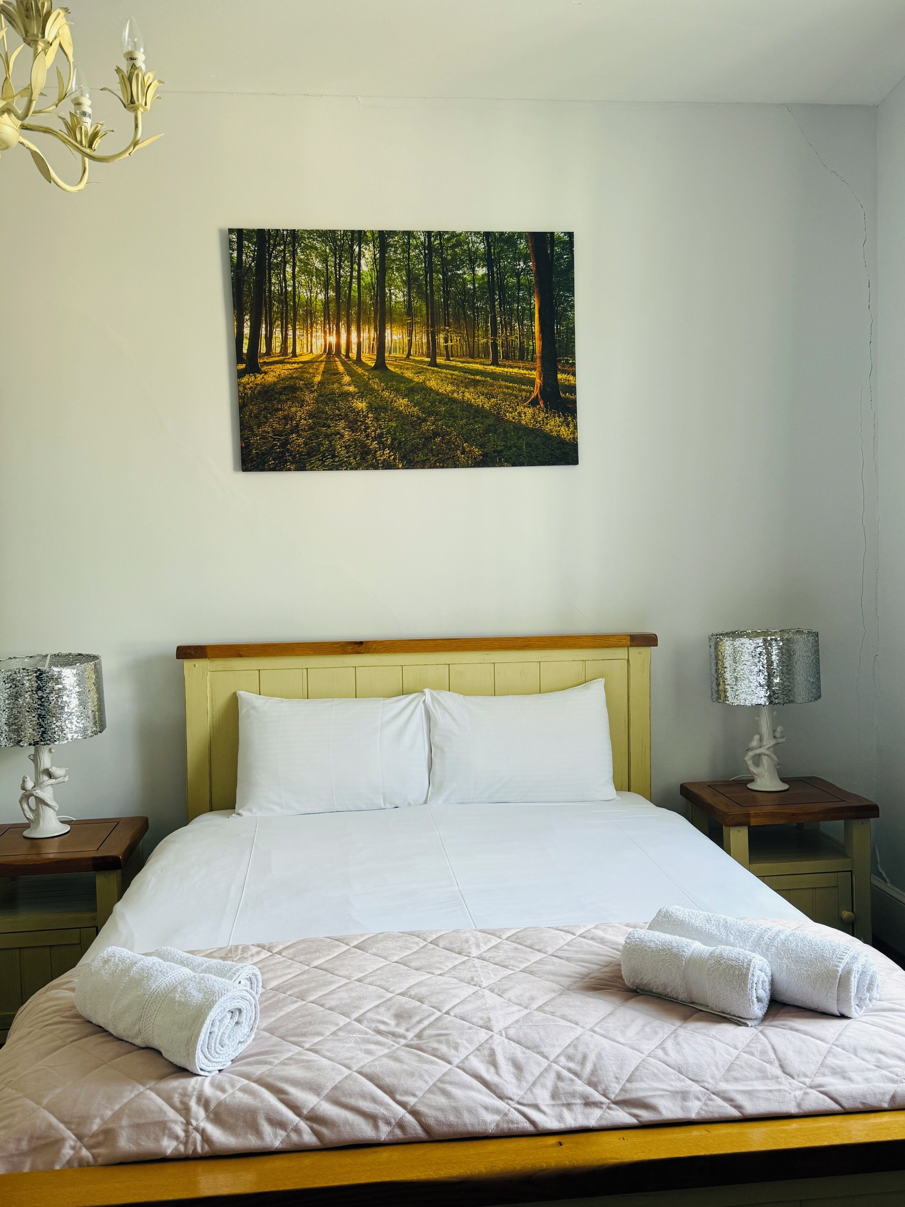 A bedroom with a bed, two nightstands, a chandelier, a painting of a forest, and a lamp on each nightstand.