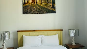 A bedroom with a bed, two nightstands, a chandelier, a painting of a forest, and a lamp on each nightstand.
