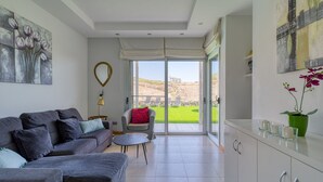 Una sala de estar moderna con un sofá gris, una mesita redonda y vista a un césped verde a través de puertas de cristal.