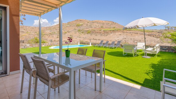 Un patio con mesa y sillas de vidrio, una piscina y vistas a las colinas.