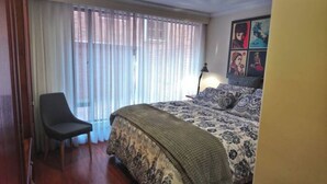 Ein Schlafzimmer mit einem Bett, einem Sessel, einem Fenster mit Vorhängen und einem Wandbild.