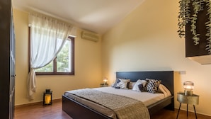 A bedroom with a bed, a window with curtains, a bedside table with a lamp, and a hanging plant.