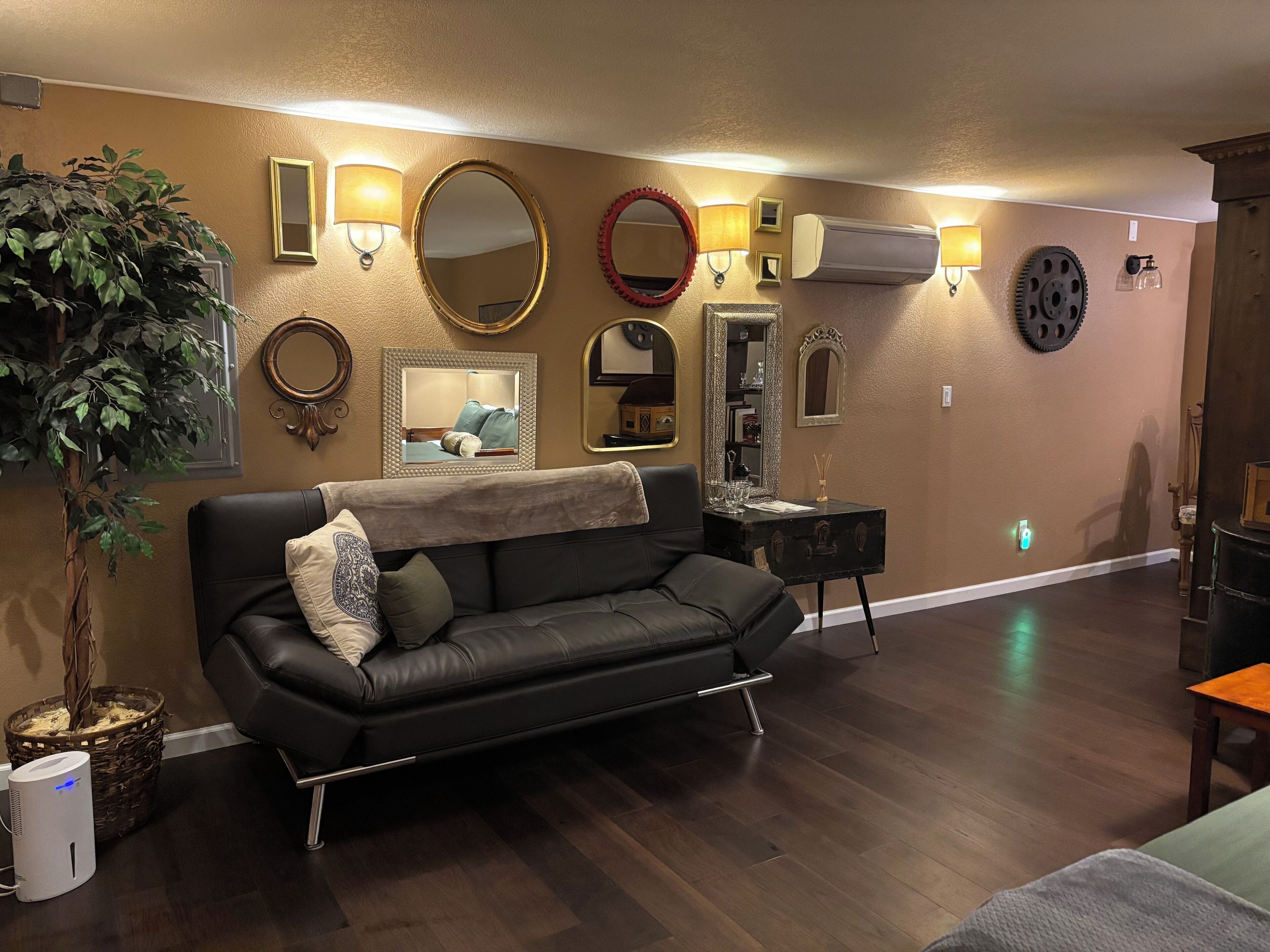 Uma sala de estar com um sofá de couro preto, uma planta em vaso e uma parede decorada com vários espelhos e arandelas.