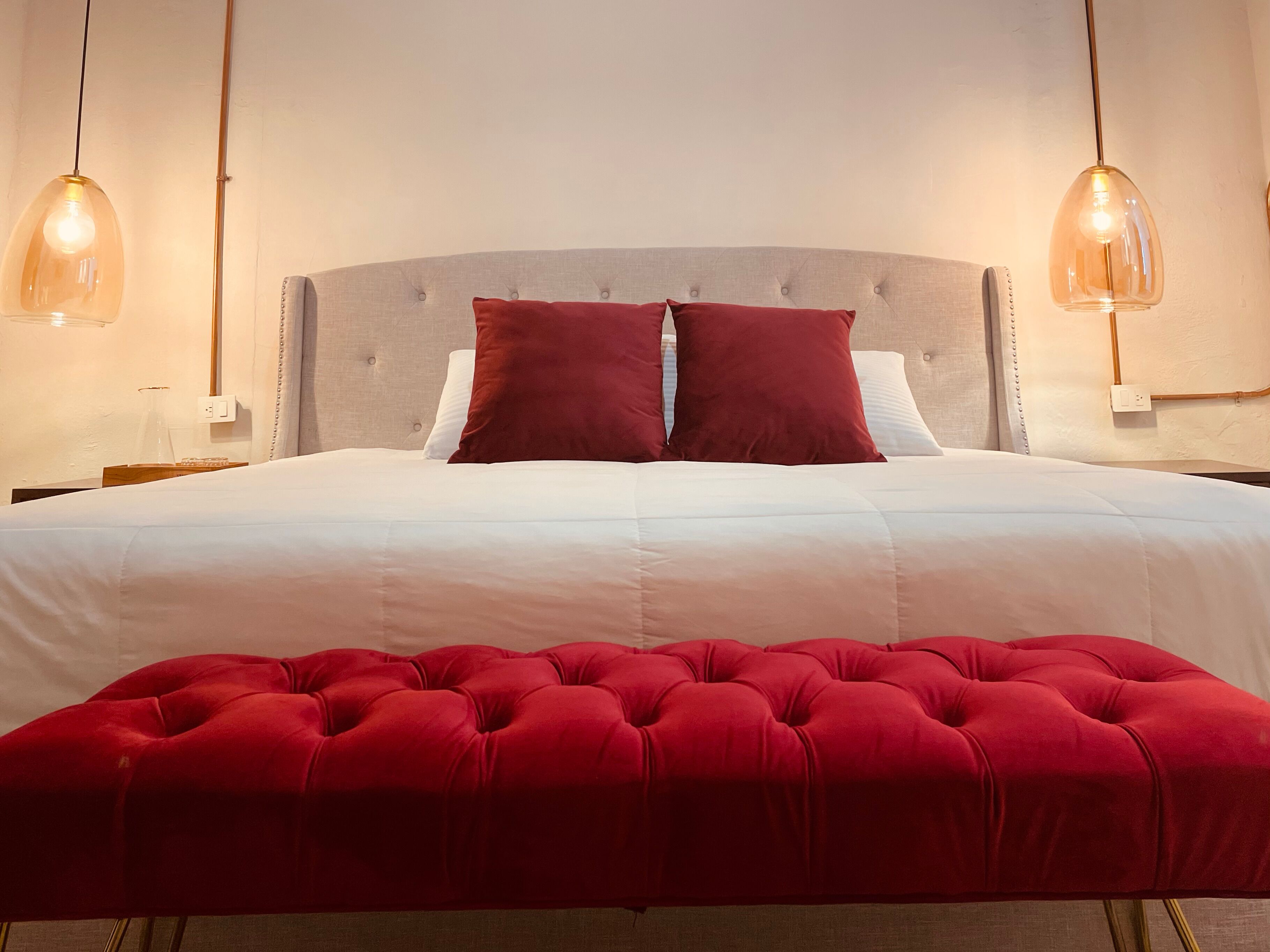 A neatly made bed with white linens and two red pillows, a tufted red ottoman in front, and pendant lighting on either side.