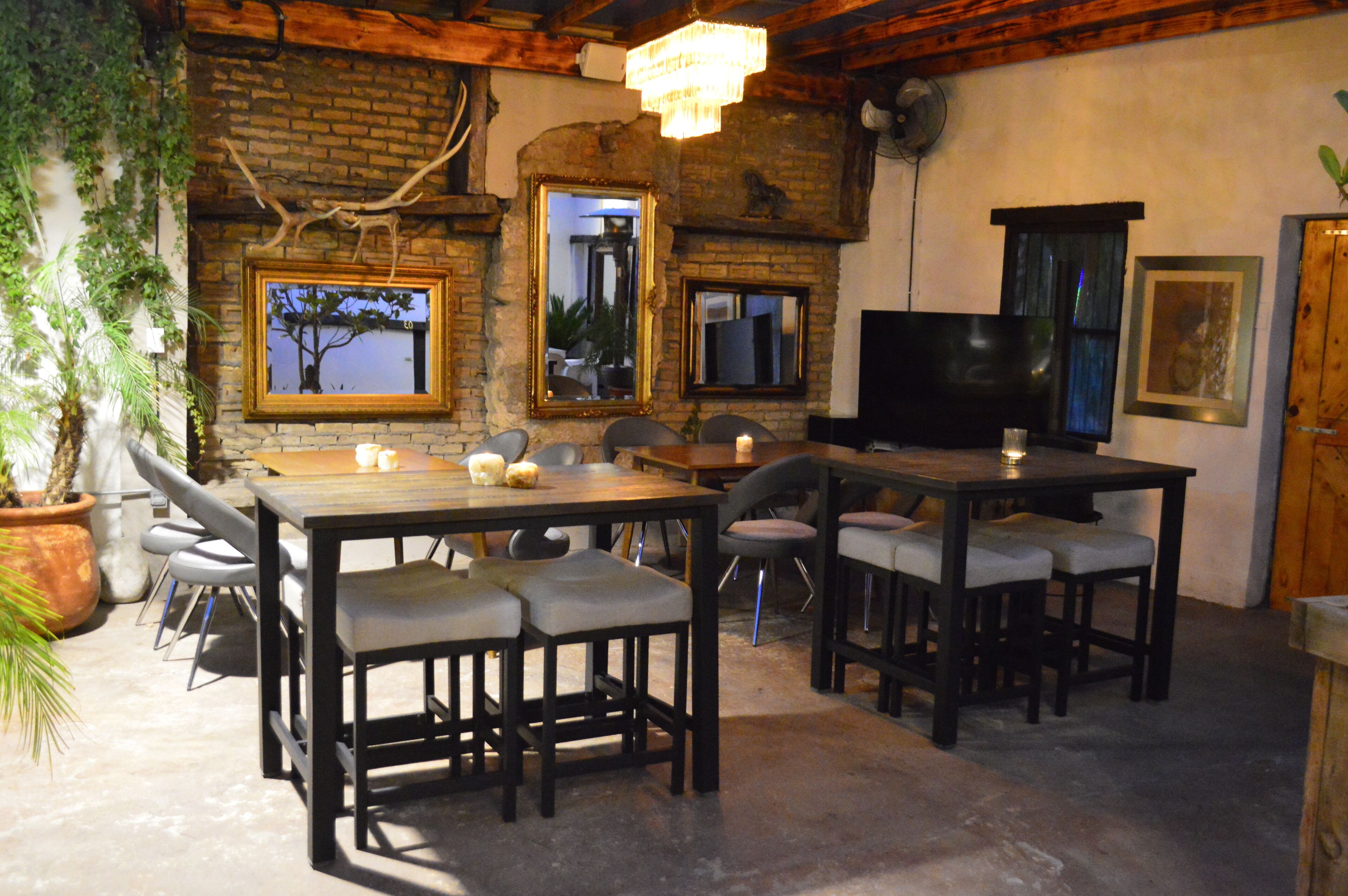 A rustic dining area with brick walls, wooden beams, and a chandelier. There are multiple tables and chairs, and a television is mounted on the wall.