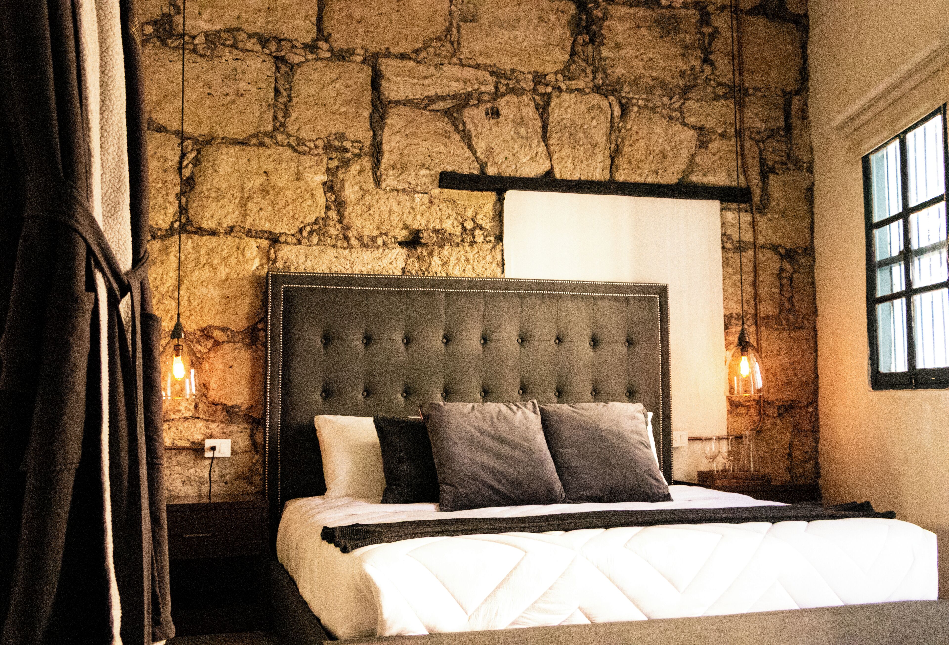 Un dormitorio con pared de piedra, cabecera capitonada y cama con ropa de cama blanca y varias almohadas.