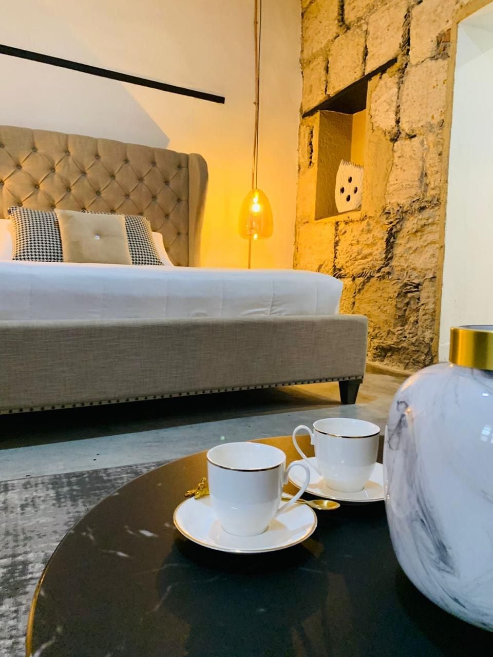 A hotel room with a tufted headboard bed, a stone accent wall, a hanging light fixture, and a table with two cups and a vase.