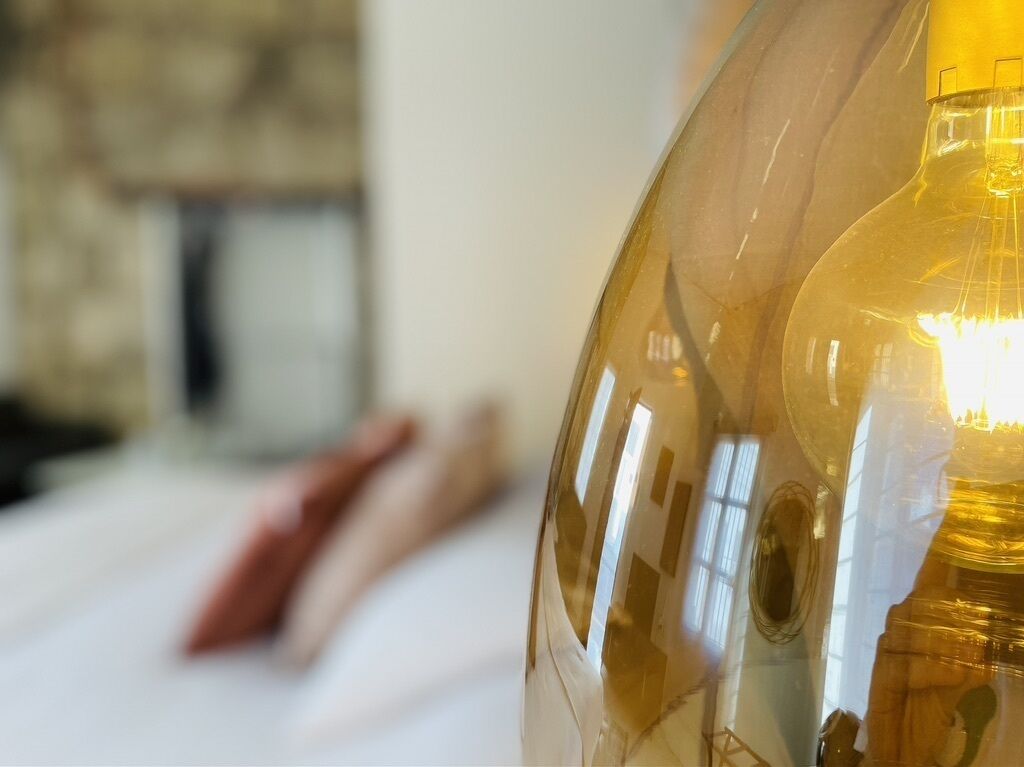 A close-up of a clear glass lamp with a warm light bulb, reflecting a room interior.