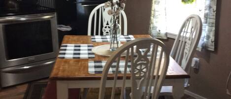 Un'area da pranzo con tovaglia a scacchi, sedie bianche, un vaso con fiori, una sveglia e un cartello con la scritta 'Farmhouse'.