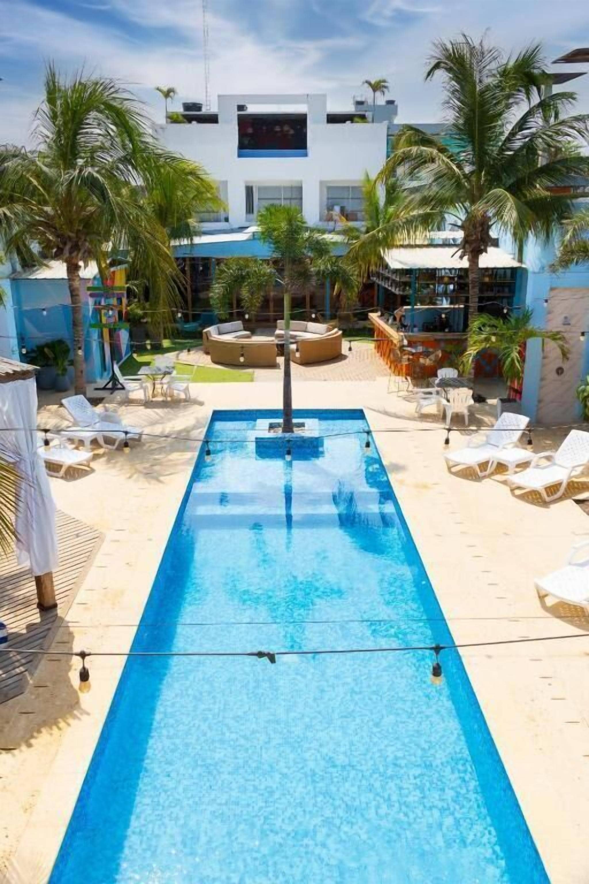 Une piscine rectangulaire, agrémentée de chaises longues et de palmiers, et entourée d’un bâtiment au design moderne.