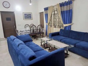 Una sala de estar con sofás azules, una mesa de centro de vidrio, una mesa de comedor con sillas y cortinas estampadas.