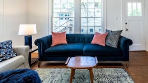 A living room with a dark blue sofa, two cushions, a small wooden table, a lamp, and a patterned rug. - The Charmer, Super Close to UT + Downtown (Austin)