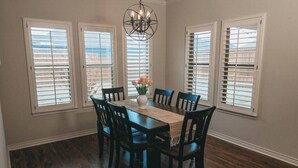 A dining room with a wooden table, six chairs, a chandelier, and three windows with blinds. - Spark Getaways: The Ricardo (Lubbock)