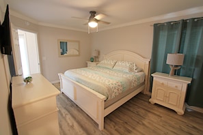 Een slaapkamer met een bed, nachtkastje, dressoir, plafondventilator en raam met gordijnen.