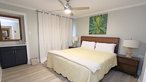 A bedroom with a large bed, a nightstand with a lamp, a ceiling fan, and a painting on the wall. - 👣 Come get your toes in the sand Call today to book your Vacation! (Gulf Shores)