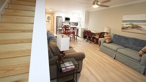 A living room with a sofa, a coffee table, and a TV. There is a kitchen in the background with a refrigerator and a microwave. - 👣 Come get your toes in the sand Call today to book your Vacation! (Gulf Shores)