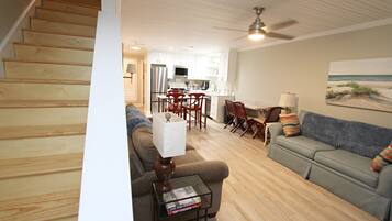 A living room with a sofa, a coffee table, and a TV. There is a kitchen in the background with a refrigerator and a microwave.