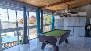 Una mesa de billar en una sala con ventanas amplias, un ventilador de techo y suelo de baldosas.