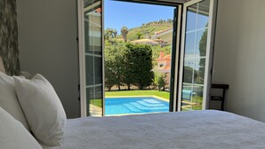 A bedroom with a large bed, a view of a swimming pool, and a hillside with houses. - Casa Roc Madeira | 5 Bedrooms | Ocean and City Views, Private Heated Pool (Funchal)