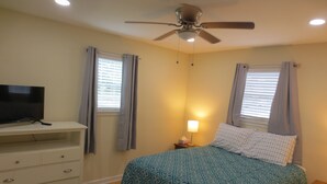 Une chambre à coucher avec un lit, un téléviseur posé sur une commode, un ventilateur de plafond et une table de chevet avec une lampe.