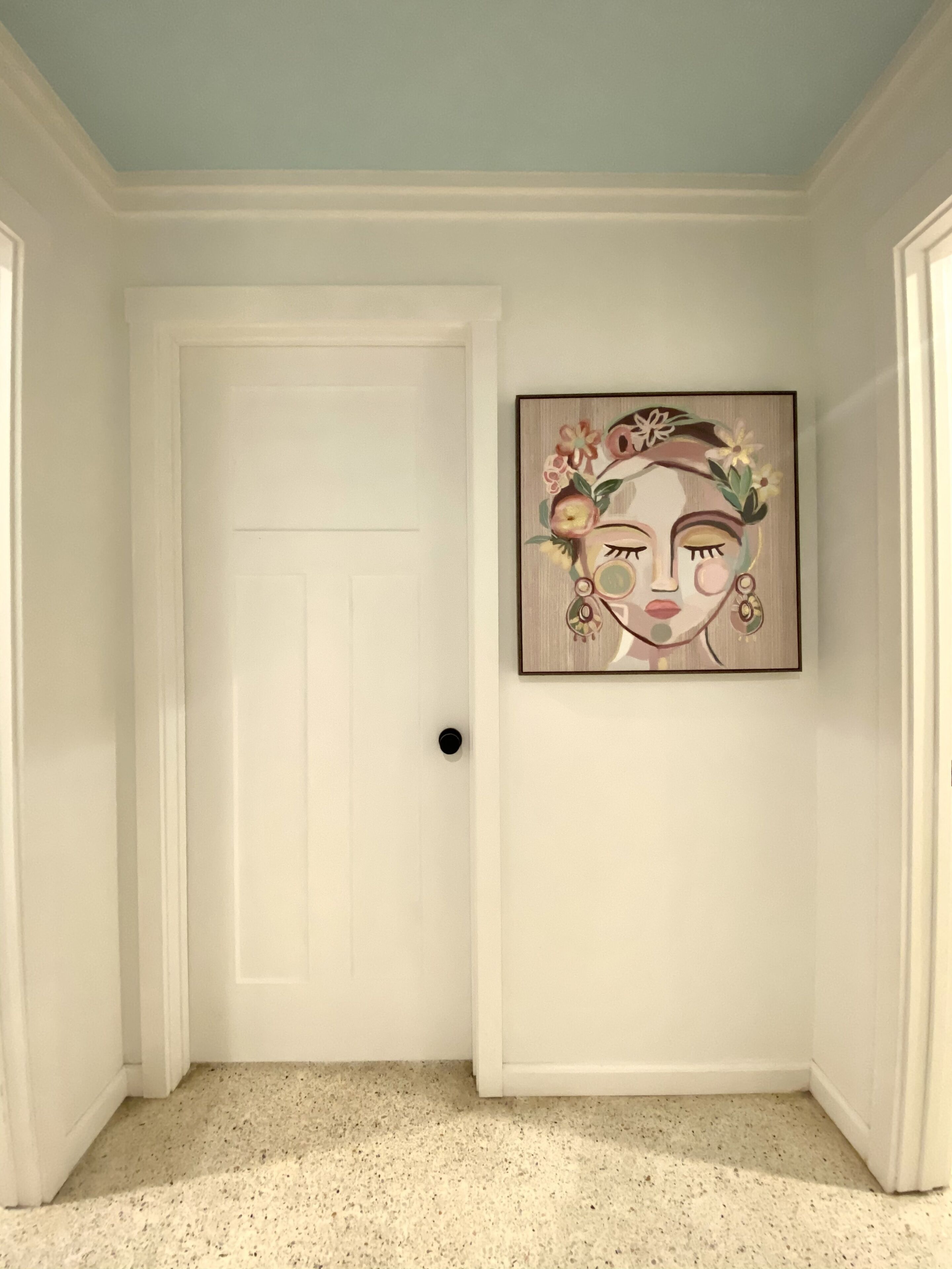 A hallway with a white door, a framed artwork on the wall, and a speckled floor.
