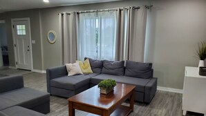 Ein modernes Wohnzimmer mit einem grauen Ecksofa, einem Holztisch und einem weißen Beistelltisch mit Pflanze.