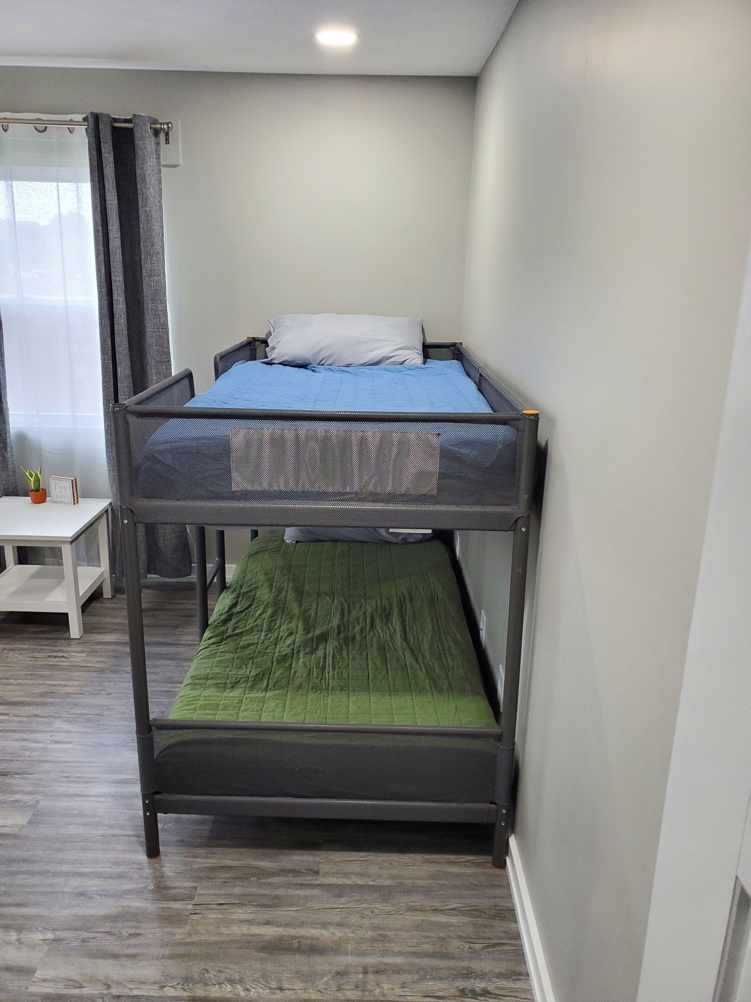 Een stapelbed met een blauwe en een groene matras, een kleine tafel met een plant en een raam met gordijnen.