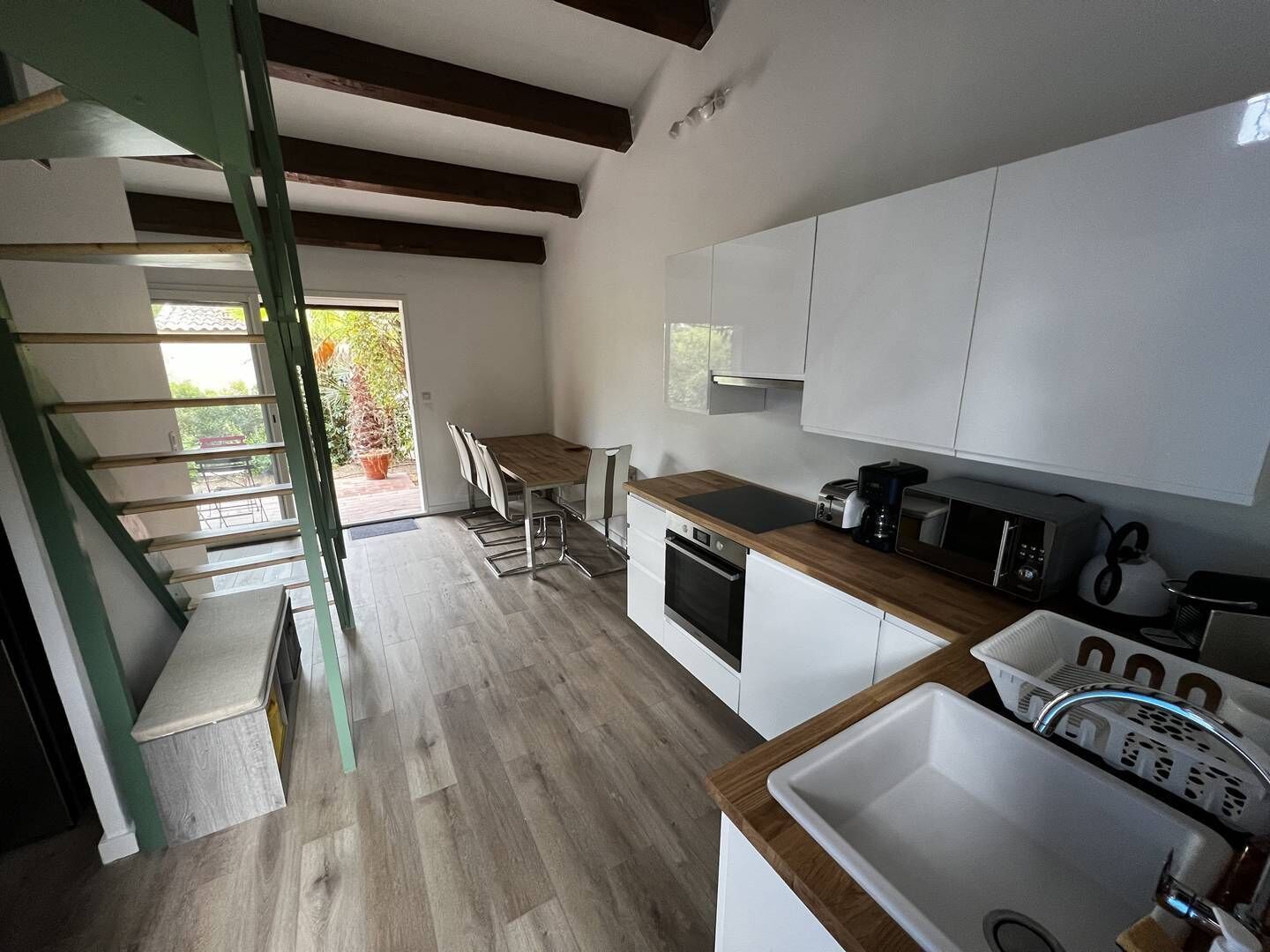 Una cocina moderna con armarios blancos, una encimera de madera y un horno empotrado. Hay un comedor con una mesa y sillas, y una escalera que lleva a un espacio exterior.