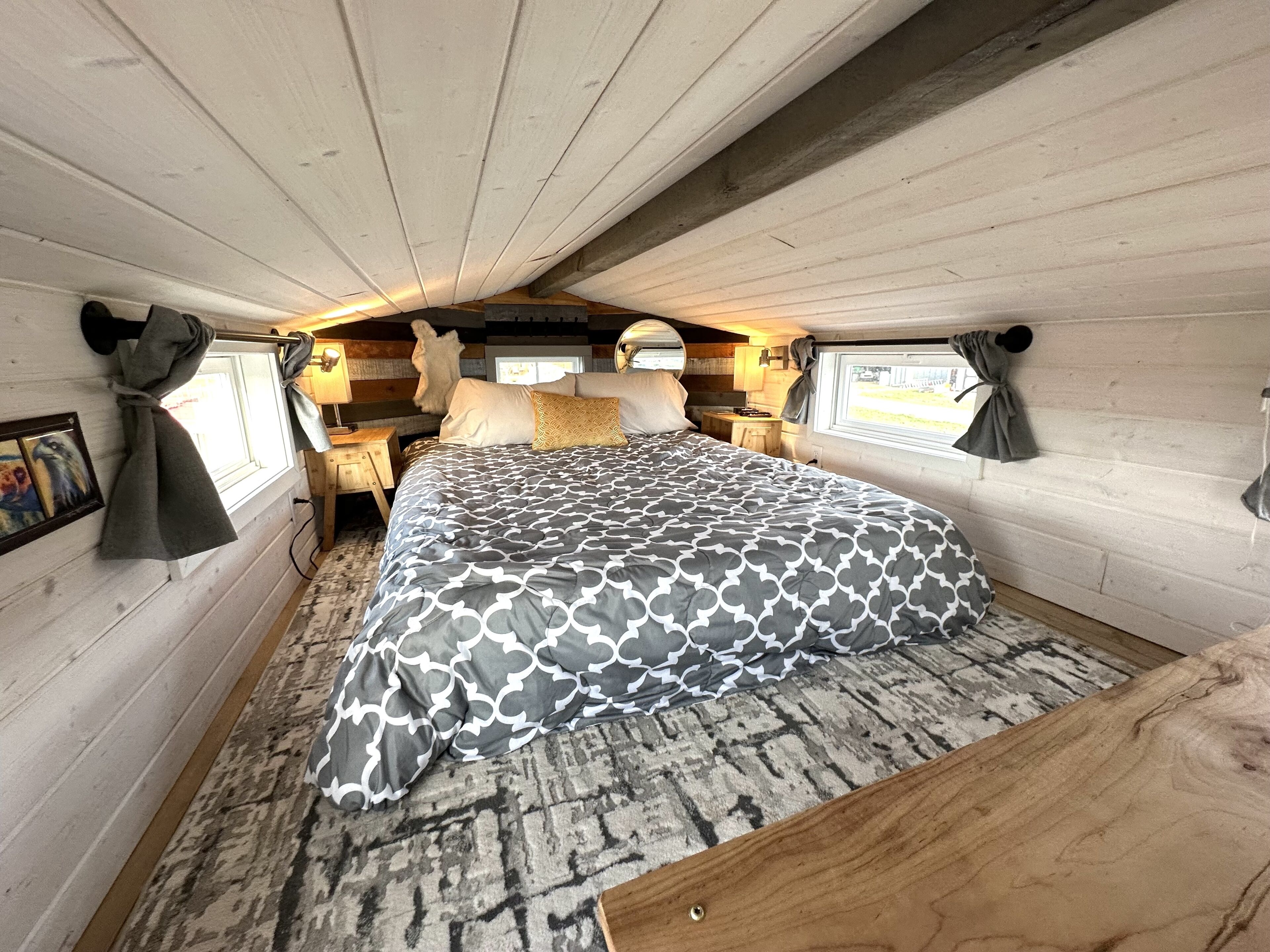 A cozy cabin bedroom with a large bed, wooden walls, a rug, and a window with curtains.