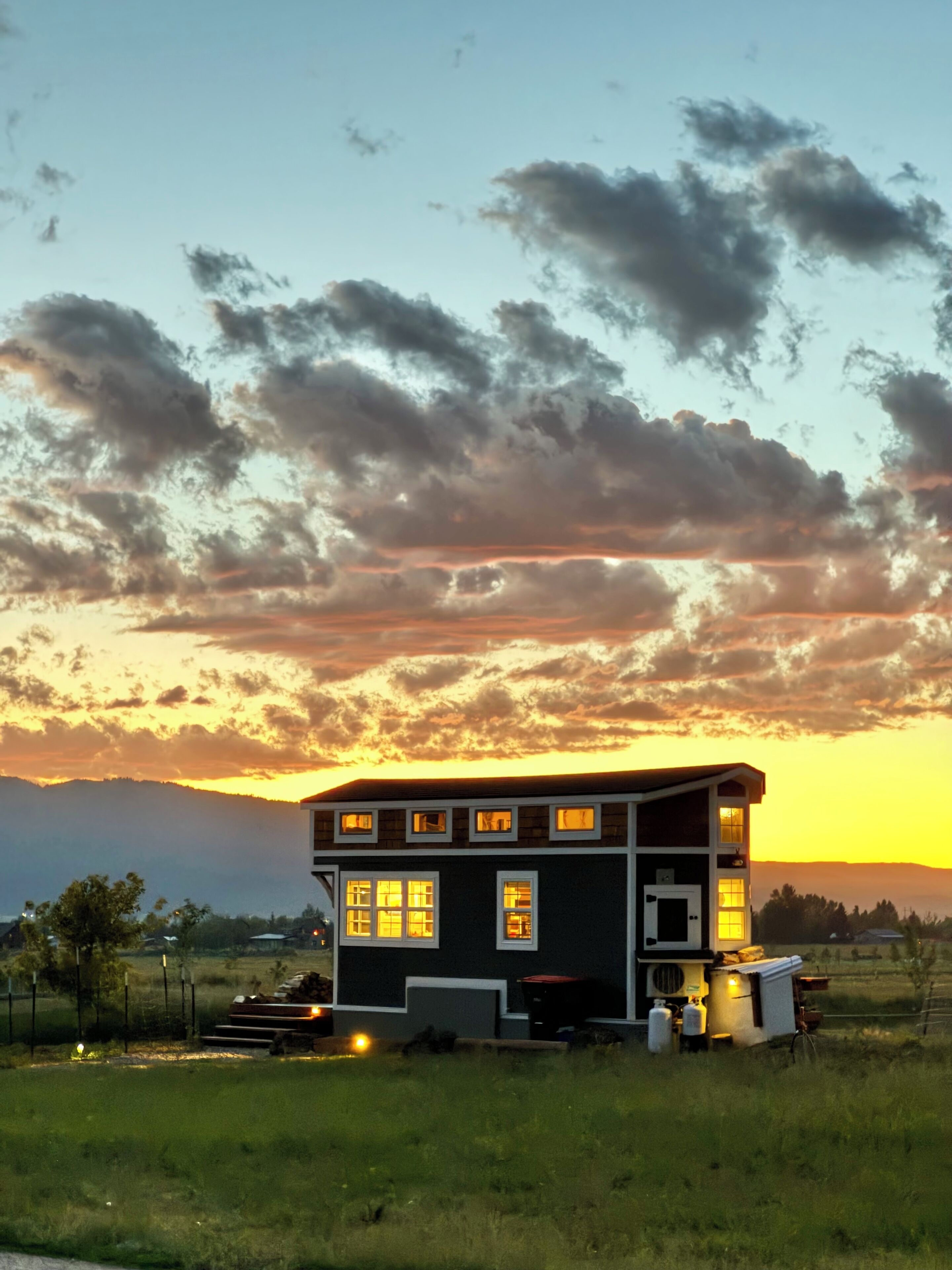 A two-story house with a dark exterior and multiple windows, surrounded by a grassy field and a mountainous backdrop during sunset.