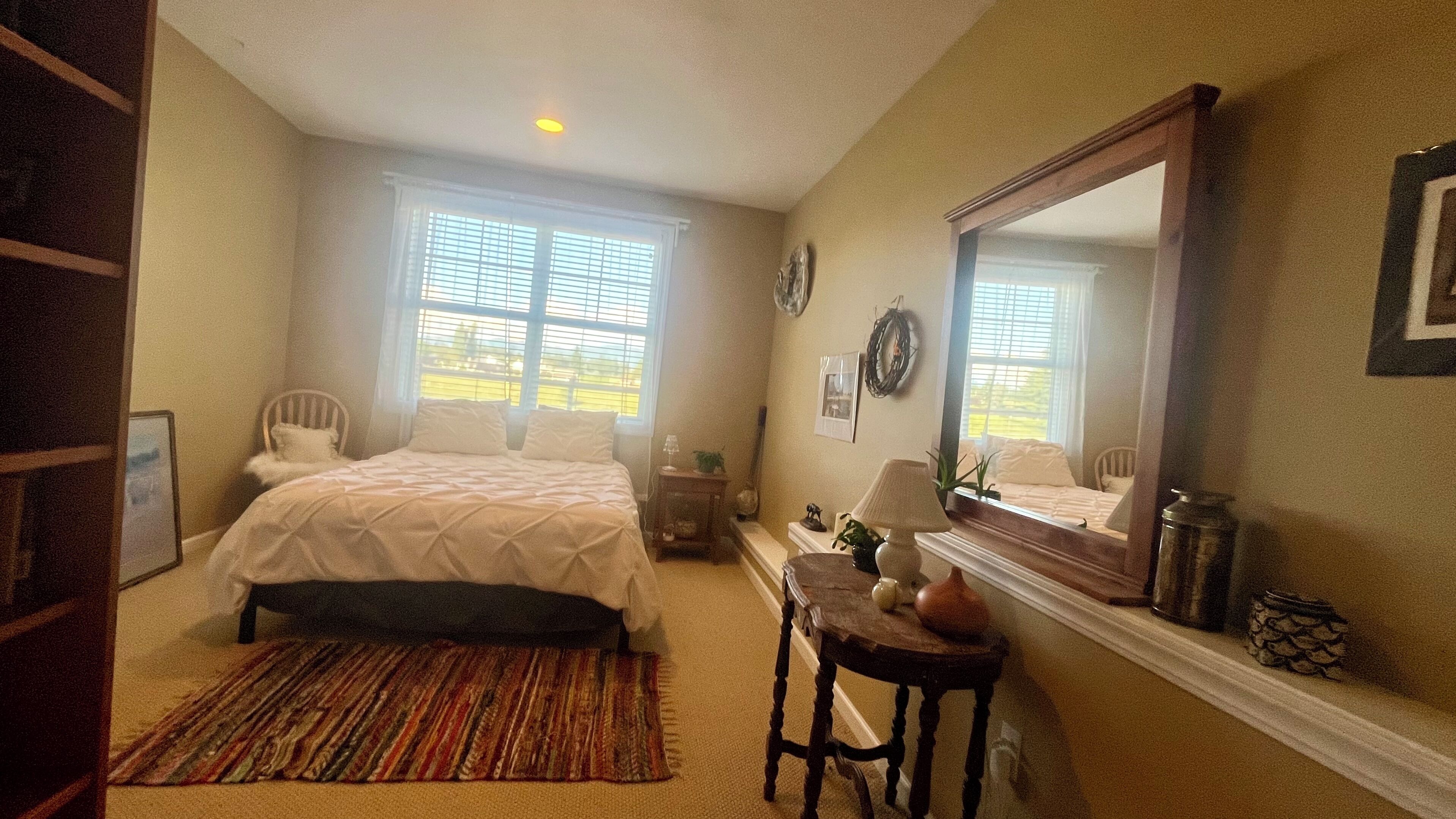 Une chambre à coucher comprenant un lit, une table de chevet, une lampe, un miroir et une vue sur l’extérieur par la fenêtre.