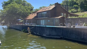 Una casa de dos pisos con piscina, rodeada de árboles y un muro de concreto.