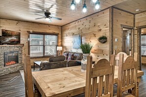 Interior acolhedor de uma cabana com lareira de pedra, mesa de jantar de madeira e ventilador de teto.