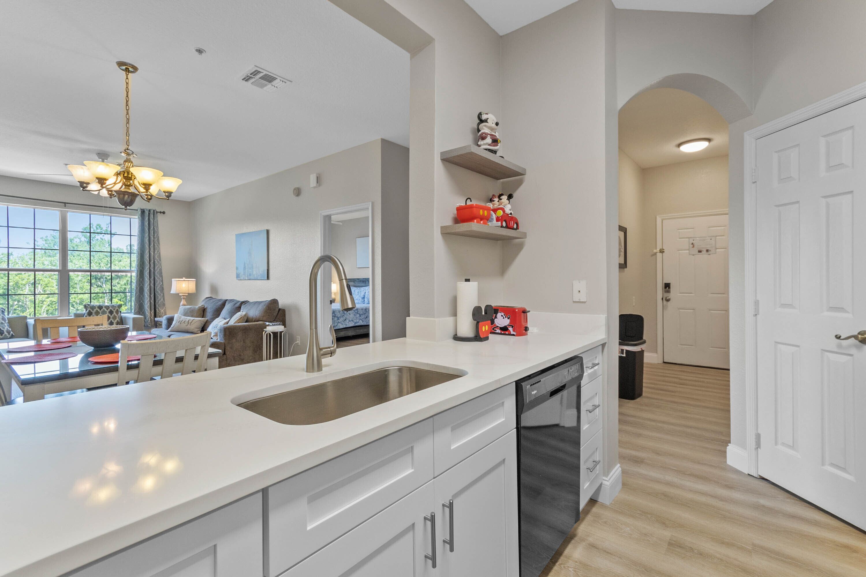A modern kitchen with a large island, stainless steel sink, and white cabinetry. There are decorative shelves with figurines and a dining area with a sofa and table.