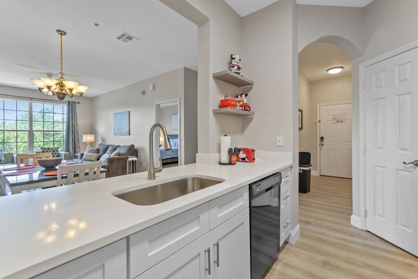 A modern kitchen with a large island, stainless steel sink, and white cabinetry. There are decorative shelves with figurines and a dining area with a sofa and table.