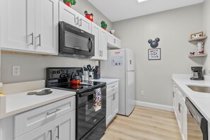 Een moderne keuken met witte kasten, een zwarte oven, een magnetron en een koelkast. Op de kasten staan decoratieve items en er is een Mickey Mouse muurdecoratie.