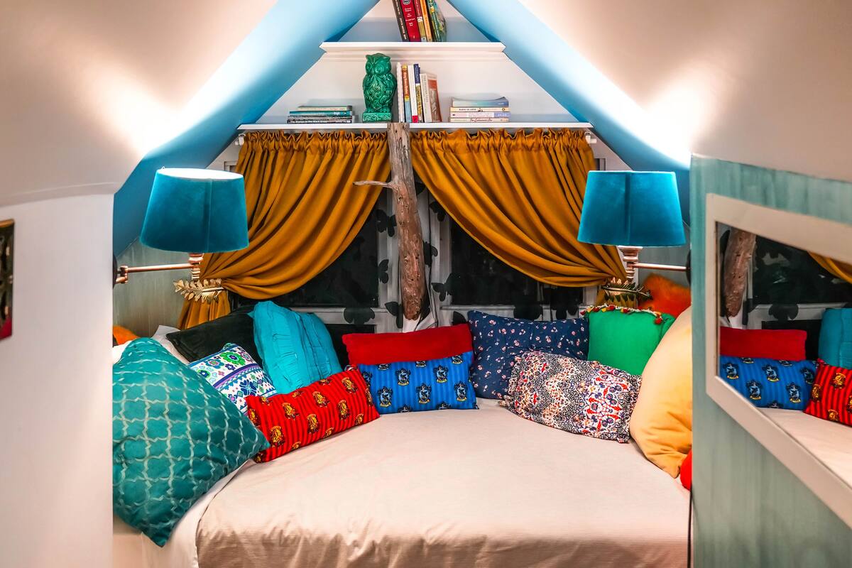 A bedroom with a bed, colorful pillows, a window with curtains, and a bookshelf.