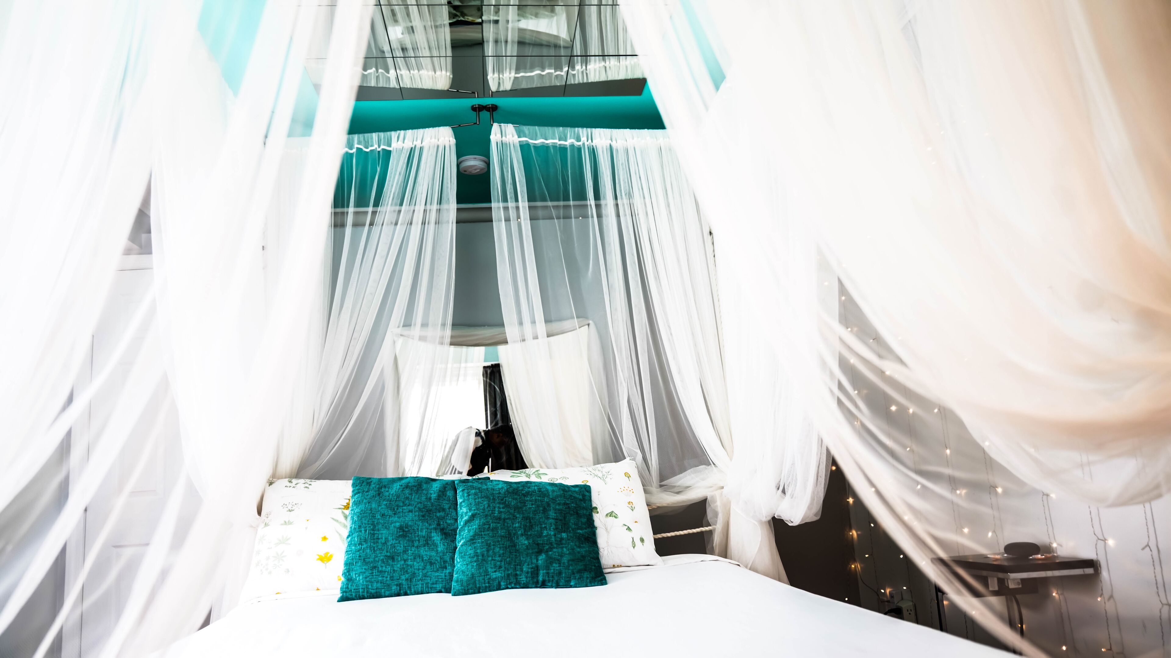Quarto com cama, almofadas azul-turquesa e cama de dossel com cortinas leves.