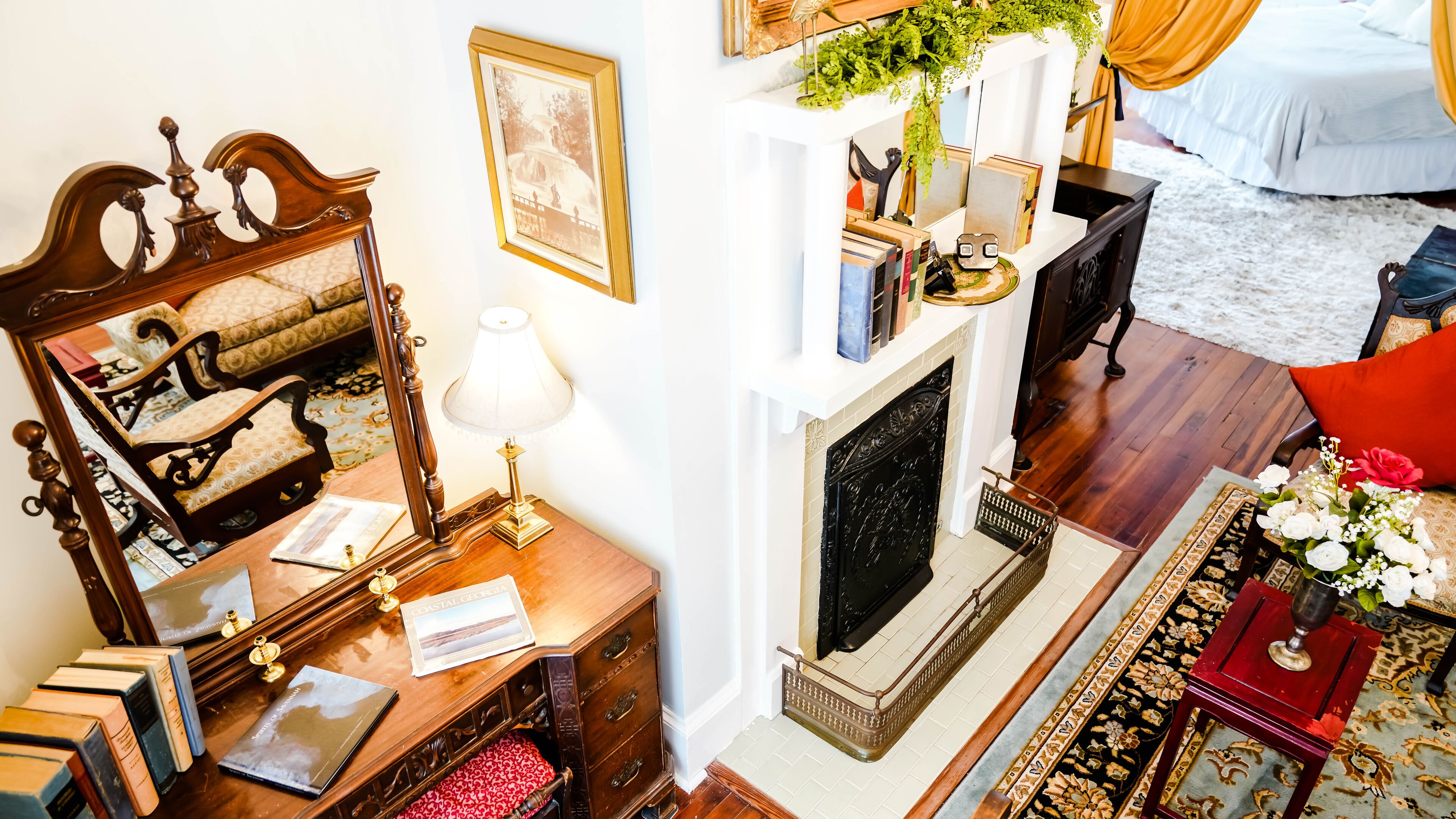 A cozy living room with a fireplace, a wooden desk with books, a mirror, and a comfortable sofa.