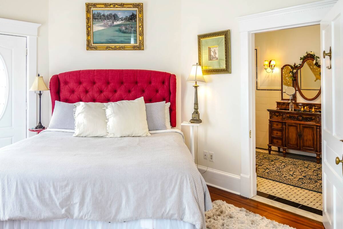 A bedroom with a red tufted headboard, a striped bedspread, and two bedside lamps. There is a door leading to a bathroom with a mirror and a vanity.