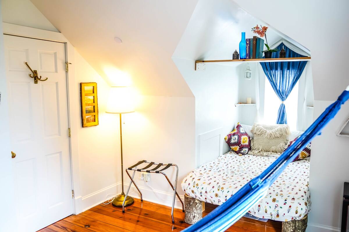 A cozy bedroom with a bed, a hanging chair, a lamp, and a shelf with books and decorative items.