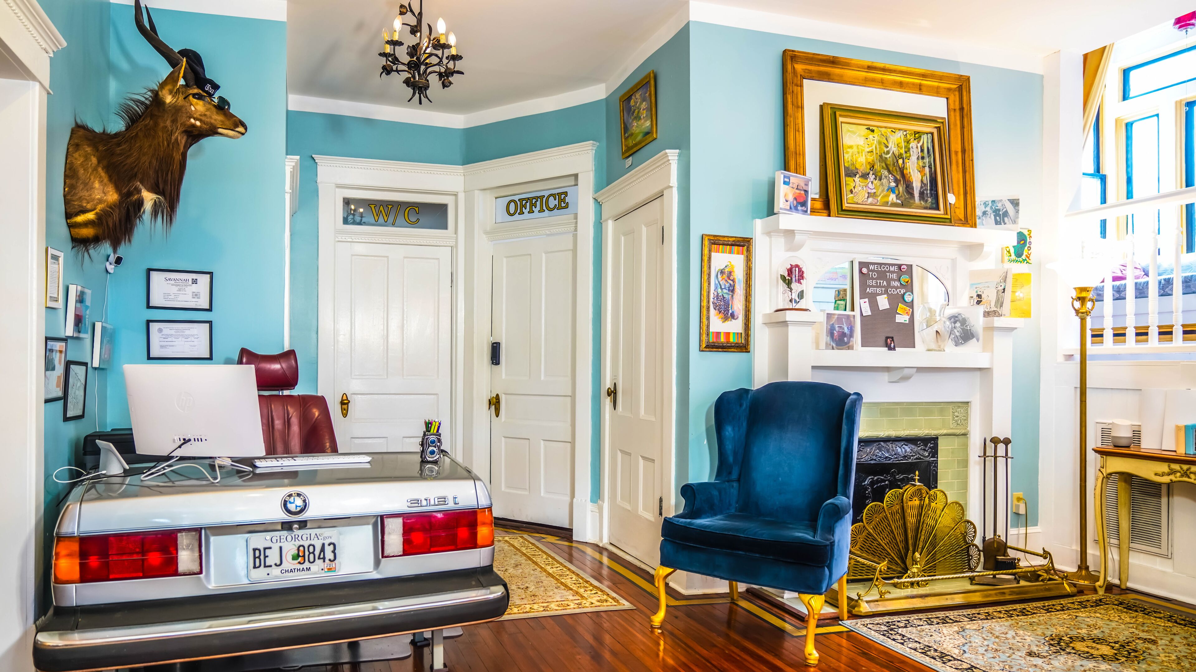 A room with a vintage car, a blue velvet chair, a mounted deer head, and a fireplace with a framed painting.
