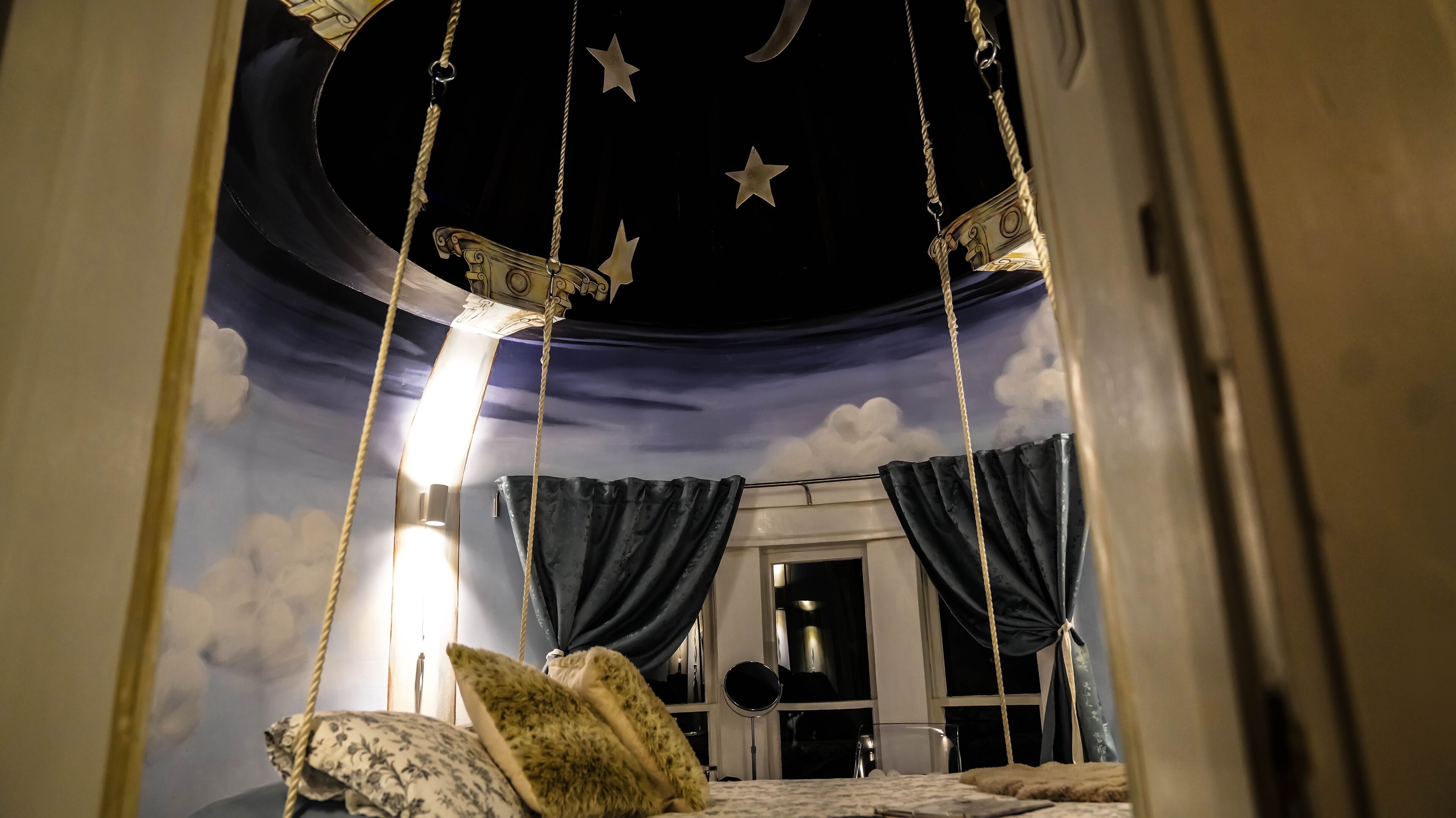 A bedroom with a starry night theme, including a hanging swing, a bed with pillows, and a night sky mural on the ceiling.