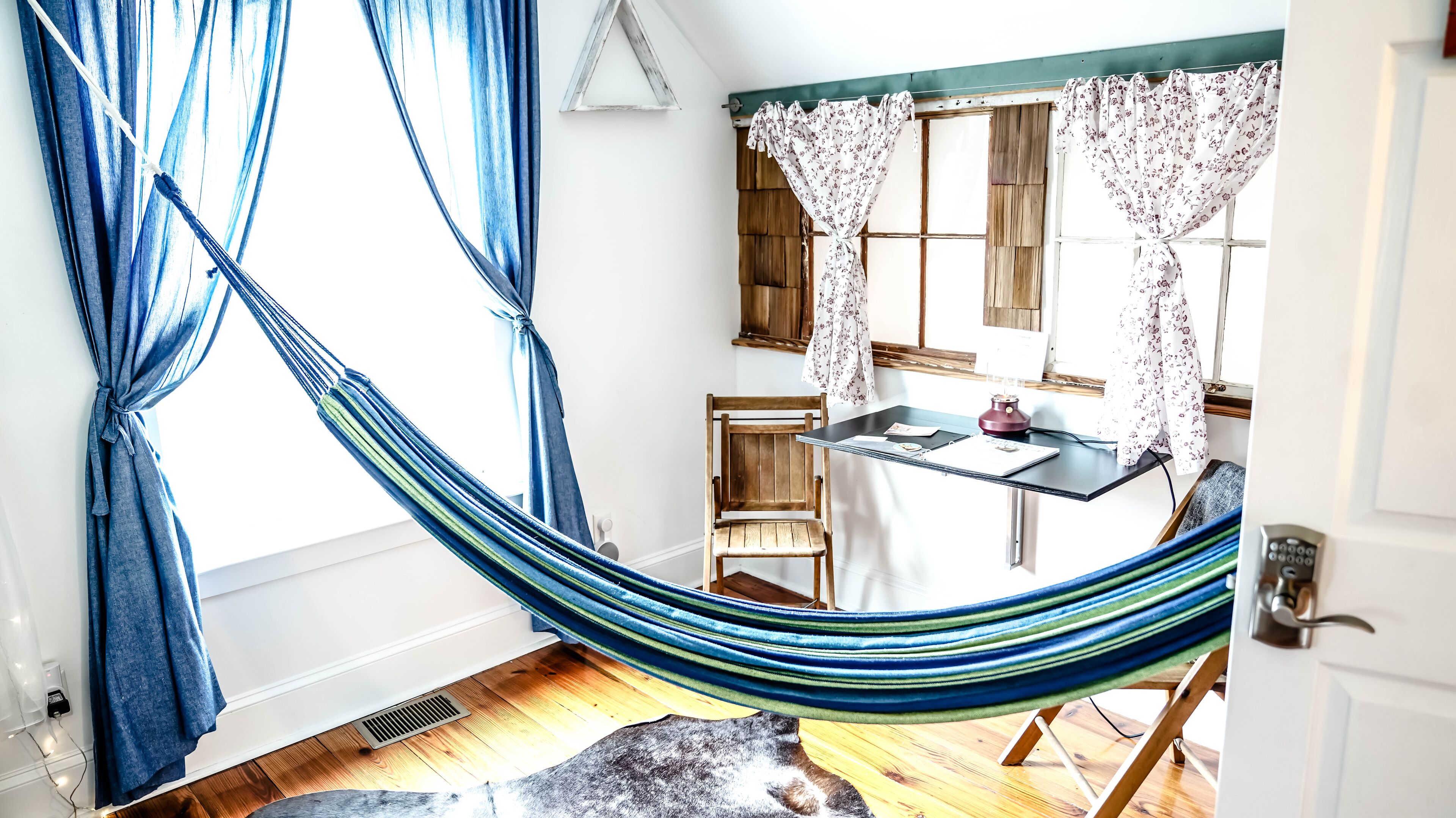 A hammock in a room with a wooden chair, a small table with a lamp, and a window with floral curtains.