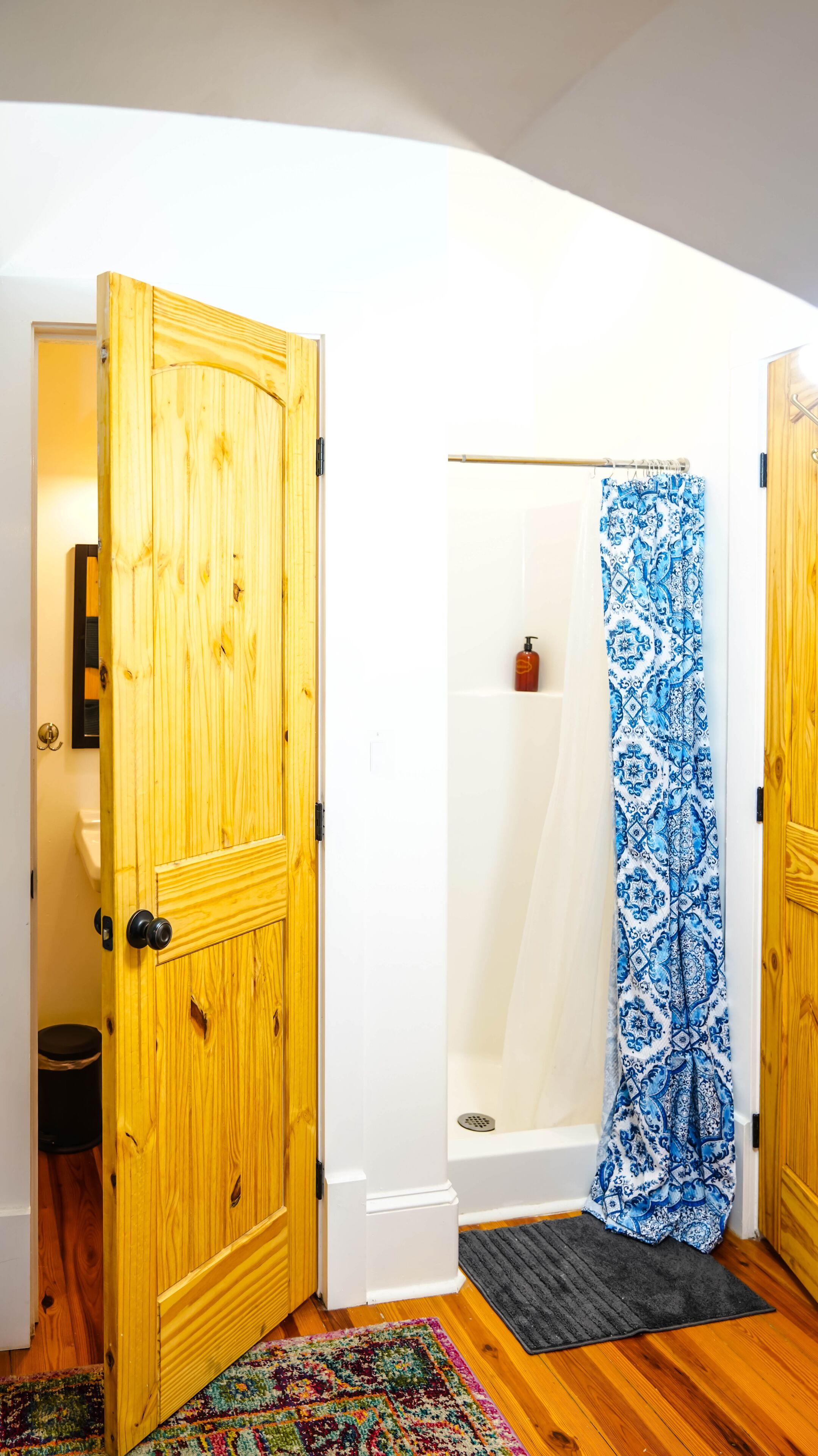 A bathroom with a wooden door, a shower with a blue patterned curtain, a white bathtub, and a colorful rug on the floor.