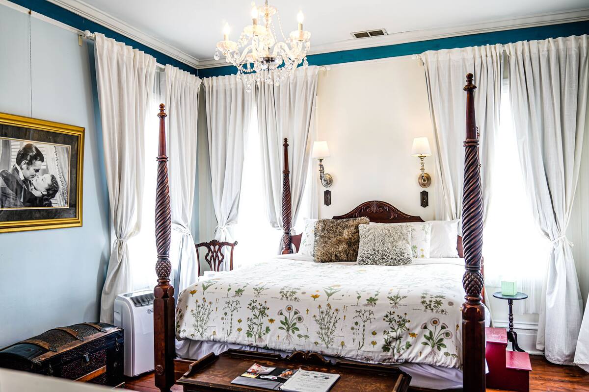A four-poster bed with a floral bedspread, a chandelier, and a framed photograph on the wall.
