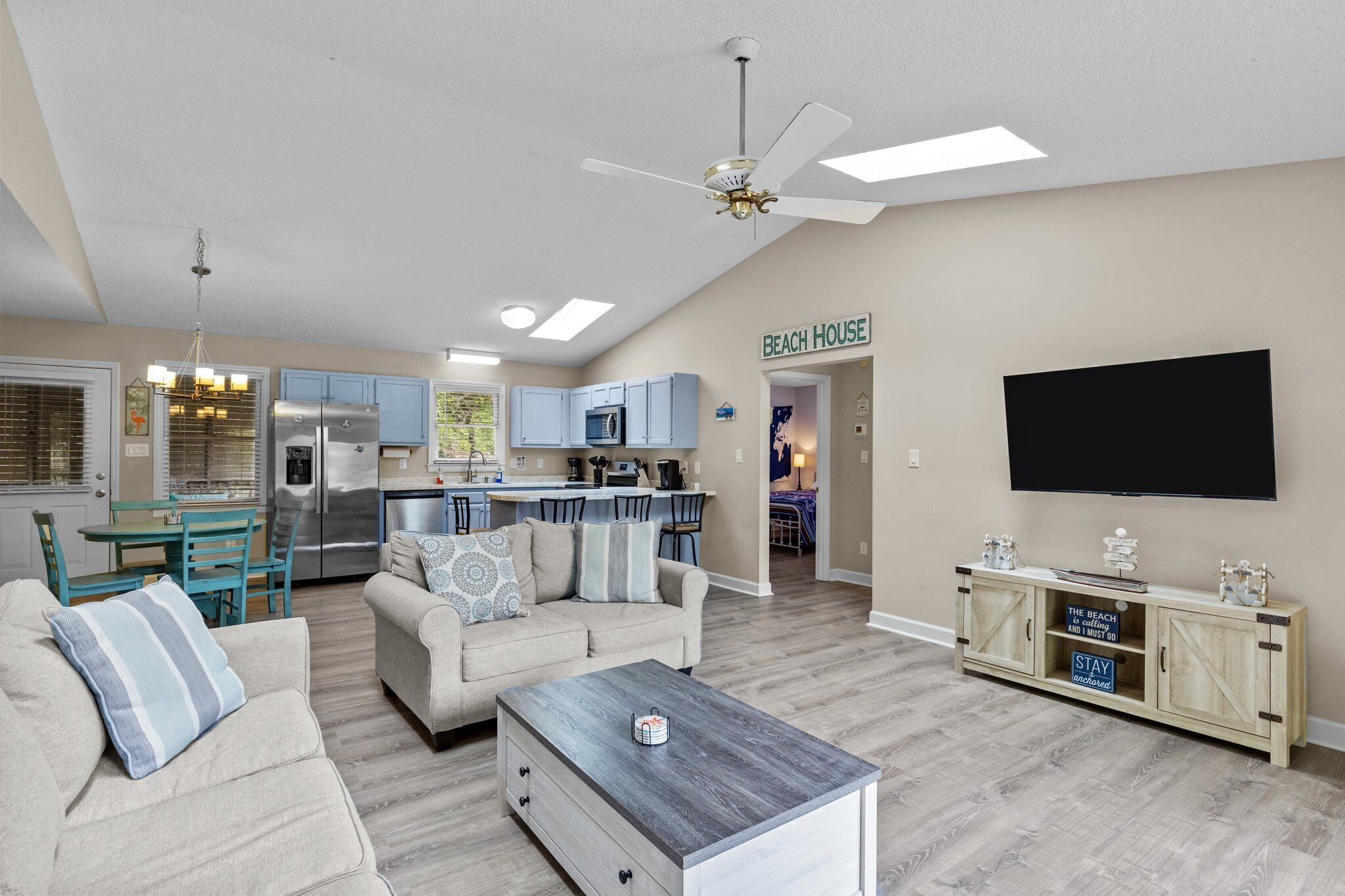 A living room with a sofa, coffee table, and TV. There's a kitchen area with a fridge, microwave, and cabinets. A sign on the wall reads "BEACH HOUSE".