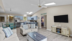 Un salón con sofá, mesa de centro y televisor. Hay una zona de cocina con refrigerador, microondas y armarios. Un letrero en la pared dice "BEACH HOUSE".